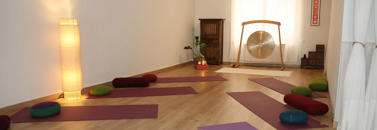 La salle de yoga : parquet clair, tapis, coussins et bolsters disposés au sol, un gong devant la fenêtre voilée de rideaux blancs